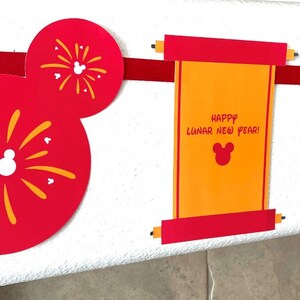 Puede incluir: Decoraciones de papel rojo y dorado para una celebraci&oacute;n del A&ntilde;o Nuevo Lunar. Las decoraciones presentan orejas de Mickey Mouse con fuegos artificiales, un rollo con el texto "&iexcl;Feliz A&ntilde;o Nuevo Lunar!" y una linterna roja con el a&ntilde;o 2025.