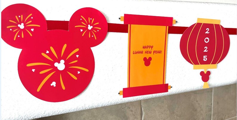 Puede incluir: Decoraciones de papel rojo y dorado para una celebraci&oacute;n del A&ntilde;o Nuevo Lunar. Las decoraciones presentan orejas de Mickey Mouse con fuegos artificiales, un rollo con el texto "&iexcl;Feliz A&ntilde;o Nuevo Lunar!" y una linterna roja con el a&ntilde;o 2025.
