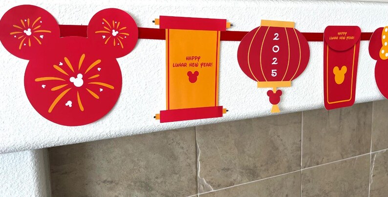 Puede incluir: Bandera roja y dorada del A&ntilde;o Nuevo Chino con orejas de Mickey Mouse, un rollo con el texto "&iexcl;Feliz A&ntilde;o Nuevo Lunar!", una linterna roja con "2025" en ella y un sobre rojo con una cabeza de Mickey Mouse.