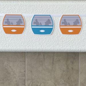May include: A colourful garland featuring nine paper cutouts of a monorail car, each with a different colour scheme. The garland is strung on a white string.
