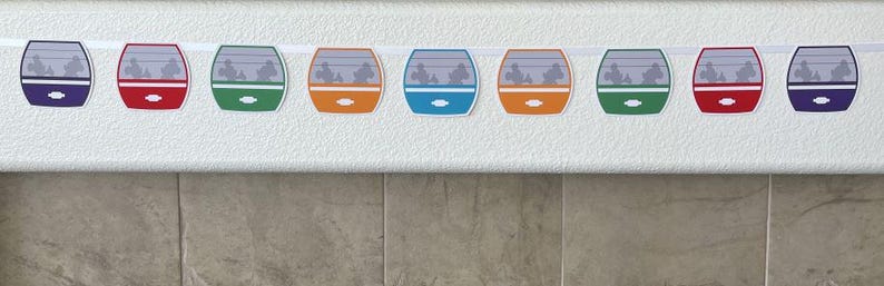 May include: A colourful garland featuring nine paper cutouts of a monorail car, each with a different colour scheme. The garland is strung on a white string.