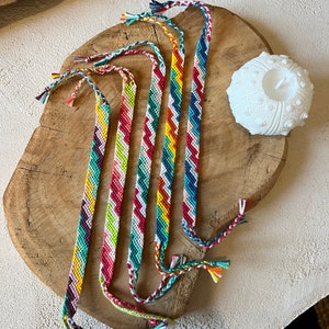 Puede incluir: Seis pulseras de amistad coloridas con patrones de chevron en tonos de rojo, amarillo, azul, verde y morado. Las pulseras están hechas con hilo de bordar y están atadas juntas en un nudo.