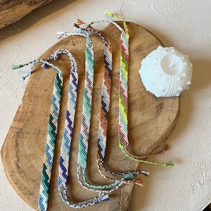 Puede incluir: Seis pulseras de amistad coloridas con patrones de chevron en tonos de azul, verde, marrón, rosa, amarillo y blanco. Las pulseras están dispuestas sobre una superficie de madera.