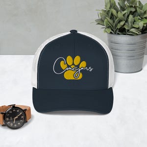 Puede incluir: Una gorra de camionero azul marino y blanca con una huella de pata amarilla y la palabra "Cougars" en escritura blanca.