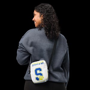 Puede incluir: Bolso bandolera blanco con un diseño amarillo y azul. El bolso tiene el texto "Cougar" en la parte delantera y una gran "S" azul con una huella de pata.