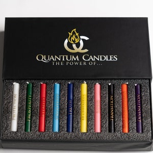Könnte beinhalten: Eine schwarze Box mit den Worten "Quantum Candles The Power Of..." auf dem Deckel. Die Box enthält 12 kleine Kerzen, jede in einer anderen Farbe und mit einem anderen Wort auf der Seite bedruckt. Die Wörter sind: Schutz, Wohlstand, Vitalität, Heilung, Wahrheit, Fülle, Liebe, Übergang, Göttlich, Erleuchtung und Manifestation.