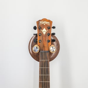 Könnte beinhalten: Eine braune Akustikgitarre mit einer Wandhalterung aus Holz. Die Gitarre hat ein goldenes Washburn X-Logo auf dem Kopf.