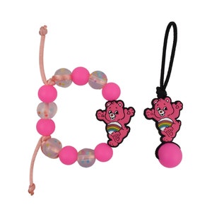 May include: Pink beaded bracelet with a pink Care Bear charm and a pink Care Bear charm on a black string.