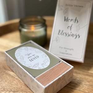 May include: A box of "Words of Blessings" cards with the text "You are so LOVED" on one card. The box is white with a floral design and a wooden drawer. A candle and a book with the text "Words of Blessings" are also visible.