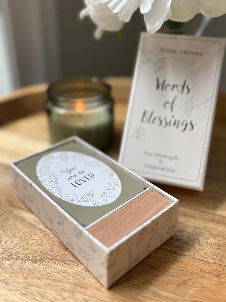 May include: A box of "Words of Blessings" cards with the text "You are so LOVED" on one card. The box is white with a floral design and a wooden drawer. A candle and a book with the text "Words of Blessings" are also visible.