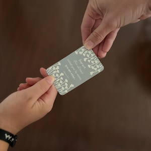 May include: A rectangular card with a light gray background and white floral accents is held between two hands. The card features the text "Fear not, For I have called you by name, you are mine." in a stylized font.