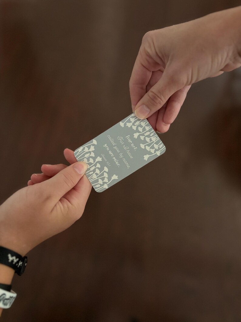 May include: A rectangular card with a light gray background and white floral accents is held between two hands. The card features the text "Fear not, For I have called you by name, you are mine." in a stylized font.