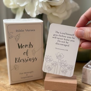 May include: A box of "Words of Blessings" Bible verses cards with floral illustrations. One card is held up, displaying a verse from Deuteronomy 31:8. The box and card are on a wooden surface, with flowers in the background.