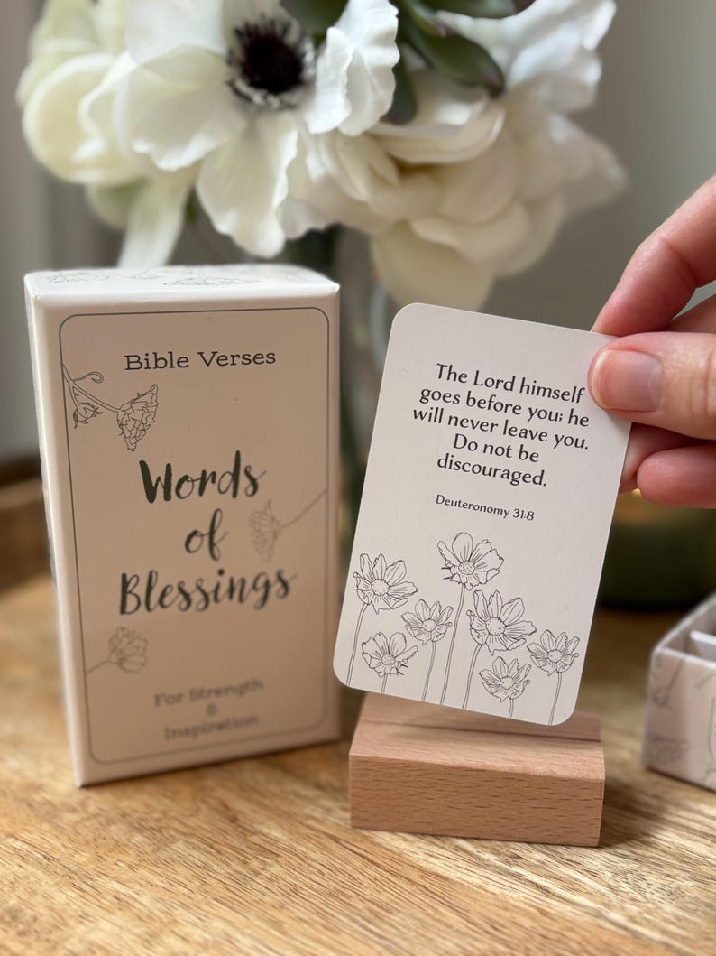 May include: A box of "Words of Blessings" Bible verses cards with floral illustrations. One card is held up, displaying a verse from Deuteronomy 31:8. The box and card are on a wooden surface, with flowers in the background.