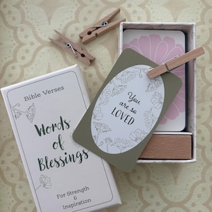 May include: A white box with the text "Bible Verses Words of Blessings For Strength & Inspiration" sits next to a box of cards. A card with the text "You are so LOVED" is held by a wooden clothespin. The background is a patterned beige surface.
