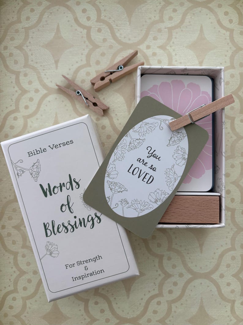 May include: A white box with the text "Bible Verses Words of Blessings For Strength & Inspiration" sits next to a box of cards. A card with the text "You are so LOVED" is held by a wooden clothespin. The background is a patterned beige surface.