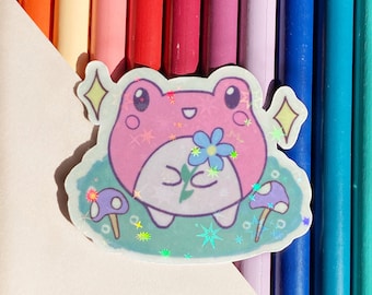 May include: A holographic sticker featuring a pink frog holding a blue flower. The frog is surrounded by green and blue glitter and two purple mushrooms.
