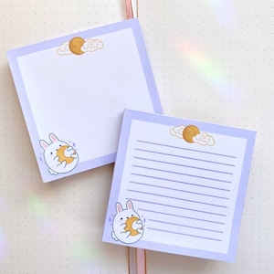 May include: Two purple lined notepads with a white bunny holding a yellow moon. The top notepad has a blank space for writing. The bottom notepad has lined paper for writing.