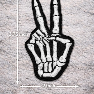 May include: Black and white embroidered patch in the shape of a skeleton hand making a peace sign. The patch is 3.75 inches tall and 2 inches wide. Text at the top reads "SKELETON PEACE SIGN" and at the bottom "IRON-ON OR SEW-ON".