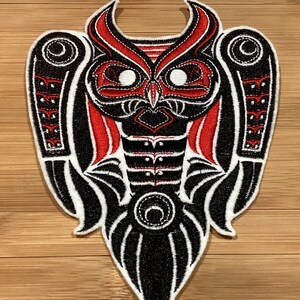 May include: Embroidered patch with an owl design in black, red, and white. The owl has a detailed feather pattern and large eyes. The patch is likely for clothing or accessories. The design is symmetrical and detailed.