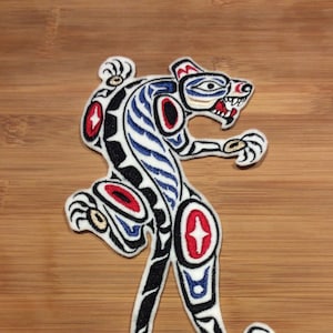 May include: Embroidered patch featuring a stylized panther design. The patch has a white background with black, red, blue, and gold accents. The panther is depicted in a dynamic pose, with its mouth open, and is likely intended for application on clothing or accessories.