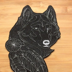 May include: A black felt patch in the shape of a wolf's head with white embroidered details. The wolf is depicted with a dreamcatcher and feathers. The patch is likely for clothing or accessories.