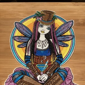 May include: Embroidered patch of a seated fairy with purple and blue wings, wearing a brown top hat and boots. The fairy is holding a blue cocktail. The background is a yellow circle with rainbow accents.