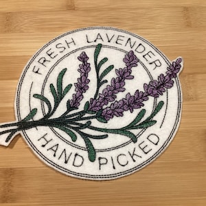May include: A white circular patch with the words "FRESH LAVENDER HAND PICKED" in black. The patch features a purple lavender sprig with green leaves. The patch is on a light brown wooden surface.