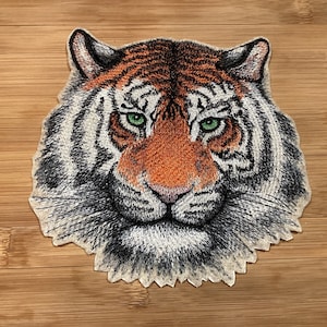 May include: Embroidered tiger head patch featuring orange, black, and white stripes, with green eyes and a pink nose. The patch is designed with detailed stitching, showcasing the tiger's facial features. This patch is suitable for jackets, bags, or other fabric items.