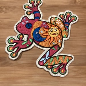 May include: Embroidered patch featuring a colorful frog design. The frog is primarily red with orange, blue, and green accents. The design includes a smiling sun, a crescent moon, stars, and a peace symbol. The patch has a cream-colored border.