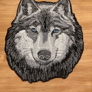 May include: Embroidered patch featuring a detailed wolf head with blue eyes. The patch is primarily black and white, with intricate stitching creating texture and depth. The wolf's expression is neutral, and the patch is set against a wooden background.