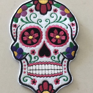 Peut inclure: Écusson de crâne de sucre brodé sur fond blanc, avec des accents floraux roses, rouges, violets et jaunes. Le crâne a des orbites noires et un nez noir. L'écusson est bordé de noir.