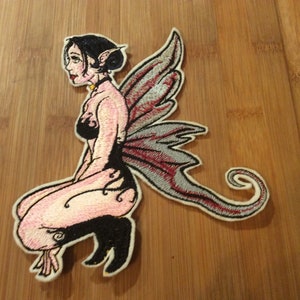 May include: Embroidered patch featuring a seated fairy with black hair, pointed ears, and large, detailed wings. The fairy is depicted in a black top and boots, with pink skin and a delicate design. The wings are gray with red accents.