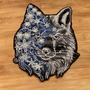 May include: Embroidered wolf patch featuring a detailed design. The left side of the wolf's face is adorned with white floral embroidery on a blue background, while the right side is in grayscale. The patch has a black border.