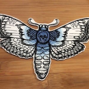 Peut inclure: Écusson brodé représentant un papillon de nuit avec un crâne. Le papillon a des ailes blanches et bleues avec des contours noirs. L'écusson est posé sur une surface en bois.