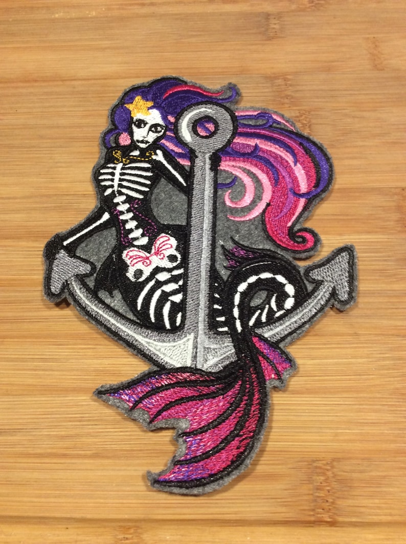 K&ouml;nnte beinhalten: Gestickter Aufn&auml;her mit einer Skelett-Meerjungfrau mit lila und rosa Haaren, einem goldenen Stern und einem schwarz-wei&szlig;en Skelett-Design. Die Meerjungfrau ist vor einem grauen Anker mit einem rosa und lila Schwanz platziert. Der Aufn&auml;her befindet sich auf einer Holzoberfl&auml;che.