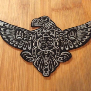 May include: A black and silver embroidered patch depicting an eagle with spread wings. The patch has detailed patterns and is suitable for application on clothing or accessories.
