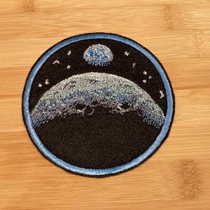 May include: Circular embroidered patch with a celestial design. A black background is framed by a blue border. The patch depicts a planet and a smaller celestial body, both in shades of blue and silver, with stars scattered around.