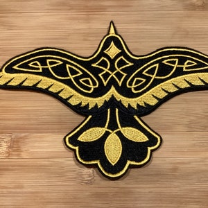Peut inclure: Écusson brodé représentant un oiseau noir stylisé aux ailes déployées. Le motif de l'oiseau intègre des entrelacs celtiques dorés et des éléments décoratifs. Idéal pour les vêtements ou accessoires.