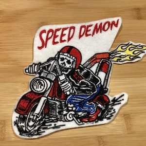 Könnte beinhalten: Gestickter Aufnäher mit einem Skelett auf einem roten Motorrad mit Flammen. Der Aufnäher trägt die Aufschrift "SPEED DEMON" in Rot auf weißem Hintergrund. Das Motorrad ist mit schwarzen und silbernen Akzenten versehen.