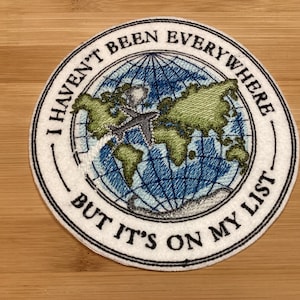May include: Embroidered patch with the text "I HAVEN'T BEEN EVERYWHERE BUT IT'S ON MY LIST". The patch features a world map with a blue and green color scheme and a gray airplane. The patch is circular with a white border.