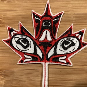 May include: Embroidered patch in the shape of a maple leaf, featuring a red, black, and white design. The design incorporates stylized shapes and patterns, with a white border. The patch is likely for clothing or accessories.