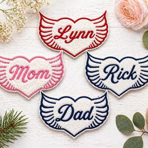 May include: Four embroidered patches in heart shapes with wing designs. The patches are white with red, pink, and navy blue accents. The patches have the names "Lynn", "Mom", "Rick", and "Dad" embroidered on them.