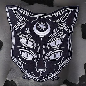 May include: Black cat patch with white embroidered detailing. The cat features four eyes and a crescent moon with a star. This patch is a unique design, ideal for adding to clothing or accessories.
