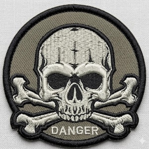 May include: Embroidered patch featuring a skull with crossbones, set against a circular olive green background with a black border. The word "DANGER" is embroidered below the skull in white letters.
