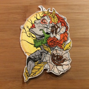 May include: Embroidered patch featuring a side profile of a woman with red hair, a green skull, and a large rose. The design includes a yellow sun-like background and gold and white accents. The patch is on a wooden surface.