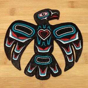 Peut inclure: Écusson brodé représentant un aigle noir stylisé avec des accents rouges, blancs et turquoise. L'aigle présente un motif en forme de cœur au centre, sur un fond en bois brun clair.