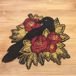 May include: Embroidered patch featuring a black bird silhouette with red roses and gold-colored leaves. The patch has a textured appearance and is likely designed to be sewn or ironed onto fabric. The design is detailed and decorative.