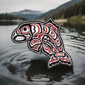 May include: A black, red, and white fish design, possibly inspired by Native American or Pacific Northwest Coast art. The fish features bold lines and complex patterns. The image is set against a backdrop of a lake and mountains.