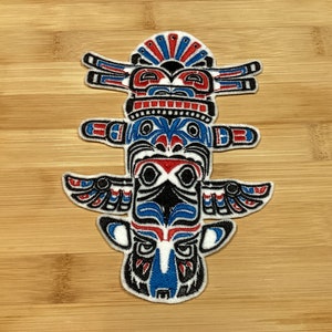 Peut inclure: Écusson brodé représentant un totem stylisé. Le patch présente une palette de couleurs vives de rouge, bleu, noir et blanc, avec des détails complexes. Le design est inspiré de l'art amérindien.